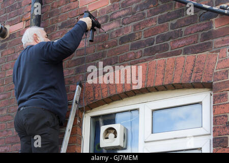 A middle-aged man standing on a ladder leaning against the exterior wall of a 1930s semi-detached house in north-west England. Stock Photo