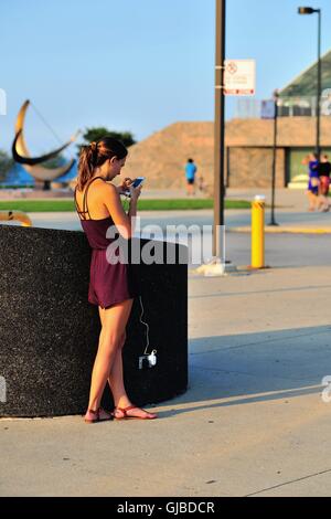 A young woman takes advantage of an outdoor electrical outlet to recharge her cell phone while also using the device. Chicago, Illinois, USA. Stock Photo