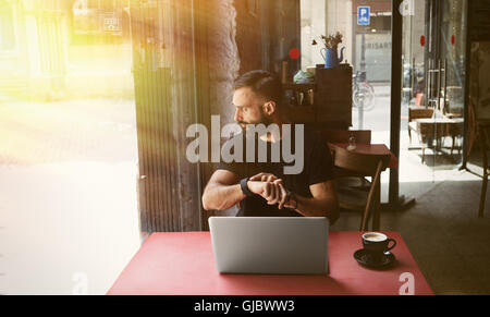 Young Bearded Businessman Wearing Black Tshirt Working Laptop Urban Cafe.Man Sitting Wood Table Cup Coffee Looking Through Window Touch Smartwatch.Coworking Process Business Startup.Blurred Background Stock Photo