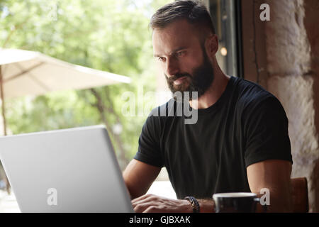 Concentrated Bearded Man Wearing Black Tshirt Working Laptop Wood Table Urban Cafe.Young Manager Work Notebook Modern Interior Design Loft Place.Coworking Process Business Startup.Color Filter.