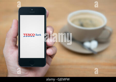 A man looks at his iPhone which displays the Tesco Bank logo, while sat with a cup of coffee (Editorial use only). Stock Photo