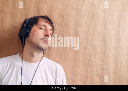 Adult male enjoying listening to favorite music podcast on headphones, man with closed eyes leaning onto wall and relaxing. Stock Photo