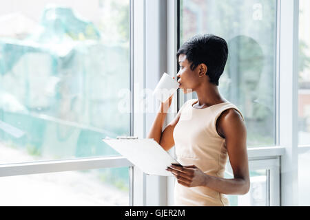 Pensive african american young businesswoman with clipboard drinking coffee and looking at the window in office Stock Photo