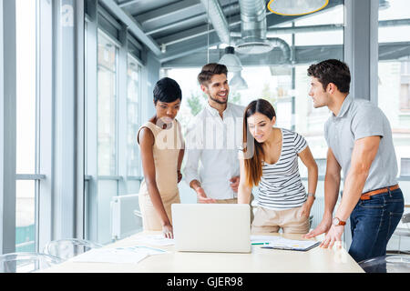 Multiethnic group of happy young business people standing and creating presentation with laptop in office Stock Photo