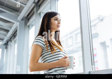 Attractive pensive young woman standing and drinking coffee near the window Stock Photo