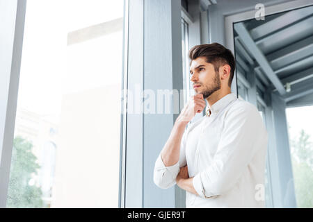 Thoughtful young man standing near the window and thinking Stock Photo