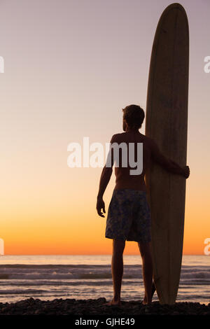 Silhouette of Man standing on beach at sunset holding longboard, San Diego, California, United States Stock Photo