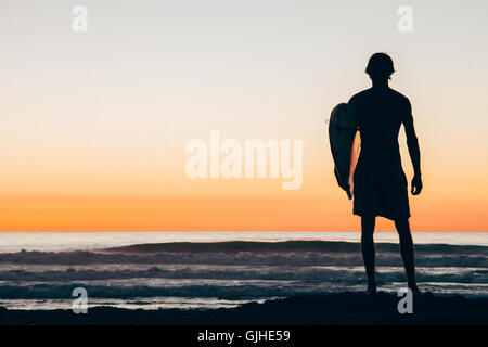 Silhouette of Man standing on beach at sunset holding surfboard, San Diego, California, United States Stock Photo