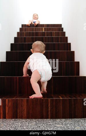 Baby boy climbing wooden stairs, sister sitting at top Stock Photo