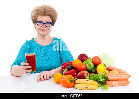 An elderly woman with glass of tomato juice in her hand sitting at table with fruits and vegetables, isolated on white backgroun Stock Photo