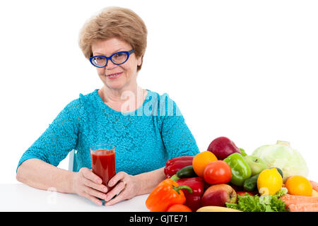 An elder woman holding glass of red tomato juice in hands, fresh fruits and vegetables are on table, isolated on white backgroun Stock Photo