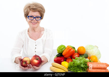 Happy senior Caucasian woman stretching hands with two big red apple, vegetables and fruits are on table, white background Stock Photo