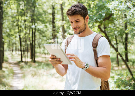 Smiling young man with backpack standing and using tablet in forest Stock Photo
