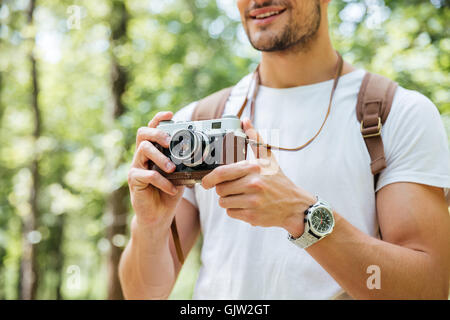 Closeup of smiling young man with backpack standing and taking photos with vintage camera outdoors Stock Photo