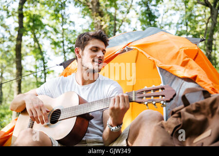 Smiling young man tourist sitting and playing guitar at touristic tent in forest Stock Photo