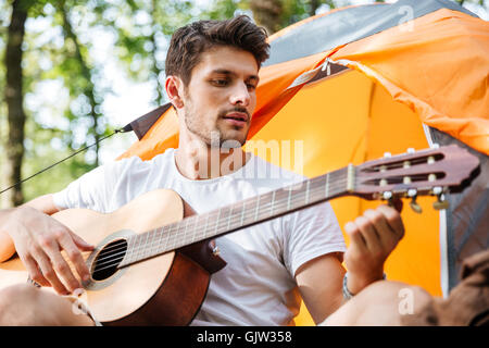 Attractive young man tourist sitting at touristic tent and playing guitar in forest Stock Photo