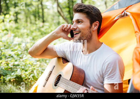 Cheerful young man tourist with guitar sitting and talking on mobile phone in forest Stock Photo