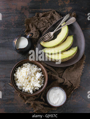 Ceramic bowl of homemade cottage cheese with yogurt jug of milk and sliced melon, served on sackcloth rag over dark textured iro Stock Photo