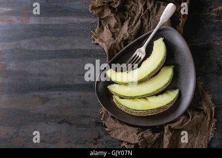 Brown ceramic triangle plate with sliced melon, served with vintage fork on wet sackcloth rag over old dark textured iron backgr Stock Photo