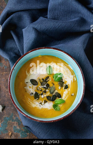 Turquoise ceramic bowl of pumpkin and sweet potato cream soup with fresh basil, cream and seeds, served on textile napkin over o Stock Photo