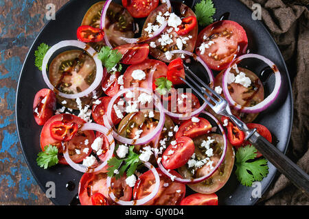 Black plate with sliced different tomatoes, red onion, balsamic sause, parsley and feta cheese salad, served with fork on wet sa Stock Photo