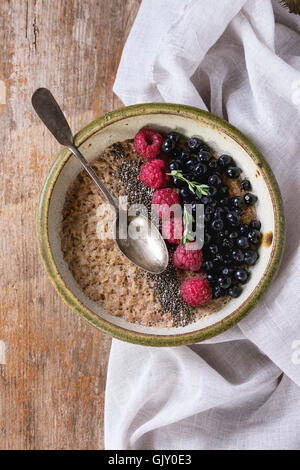 Bowl of oatmeal porridge with blueberries, raspberries and chia seeds, served with linen rag and vintage spoon over old wood tex Stock Photo