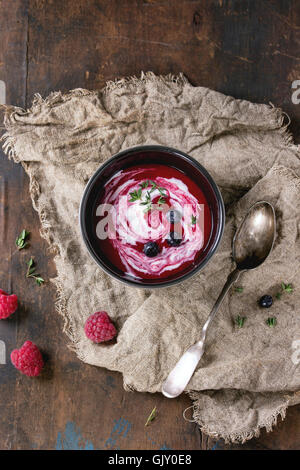 Black ceramic bowl of raspberry dessert soup with cream, served with spoon, fresh berries and thyme on sackcloth over old wood t Stock Photo