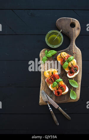 Fresh homemade toasts with mozarella, cherry tomatoes and basil served with pesto sauce on rustic wooden board over dark wooden Stock Photo