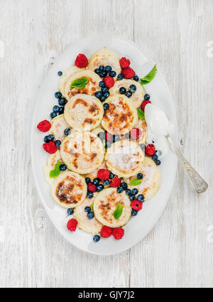 Syrniki or cottage cheese pancakes with fresh forest berries and sour cream sauce in serving dish over white wooden background, Stock Photo