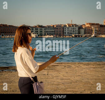 Asian woman taking a selfie with her smartphone and selfie stick by the water in Venice, making a V or peace symbol with her hand Stock Photo