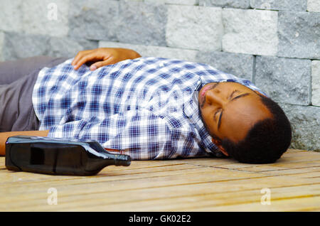 Man wearing casual clothes lying drunk passed out on wooden surface next to grey brick wall, empty bottle beside him Stock Photo