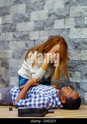 Man wearing casual clothes lying drunk passed out on wooden surface, pretty woman sitting beside him trying to get contact by touching and shaking Stock Photo