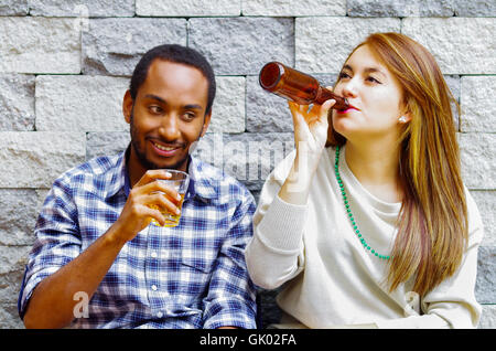 Interracial couple wearing casual clothes sitting towards grey brick wall enjoying some drinks and each others company Stock Photo
