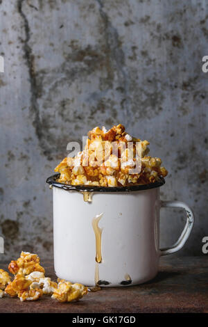 Caramelized sweet popcorn served in vintage enameled white mug with pouring caramel over old dark iron rusty background. Stock Photo