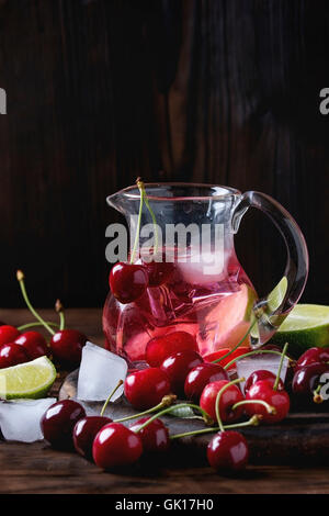 Homemade cherry and lime lemonade, served in glass jug with ice cubes, fresh cherries and sliced lime on wooden chopping board o Stock Photo