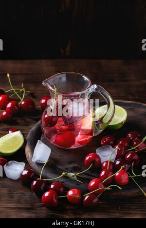 Homemade cherry and lime lemonade, served in glass jug with ice cubes, fresh cherries and sliced lime on wooden chopping board o Stock Photo