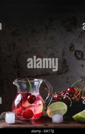 Homemade cherry and lime lemonade, served in glass jug with ice cubes, fresh cherries and sliced lime oveer dark iron rusty back Stock Photo