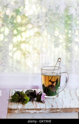Glass cup of hot herbal tea with bunch of fresh violet basil, served with vintage tea-strainer on old wooden stool with window a Stock Photo