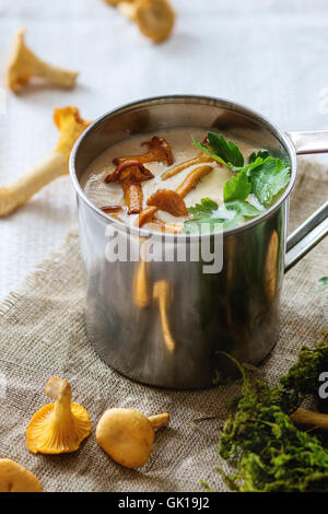 Chanterelle Cream soup in iron mug with parsley, served with fresh mushroom and forest moss over rag on white linen.Natural day Stock Photo