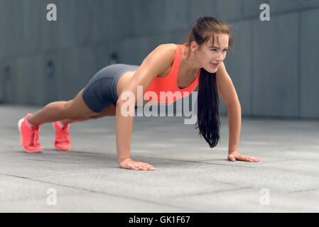 Fit young woman doing press-us in town in her sportswear looking up at the camera with a smile, frontal low angle view Stock Photo