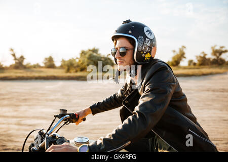 Young brutal man in a black jacket and glasses driving retro motorcycle Stock Photo