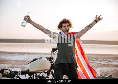 Smiling brutal man in american flag cape sitting on his motocycle with hands up in the air Stock Photo