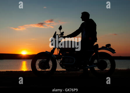 Motorbiker motorcyclist in silhouette at sunset man sitting on motorbike by a lake  one hand on handlebar Stock Photo