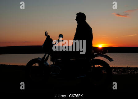 Motorbiker motorcyclist in silhouette at sunset man sitting on motorbike near water Stock Photo