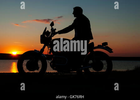 Motorbiker Motorcyclist in silhouette at sunset by lake man on motorbike one hand on handlebar Stock Photo