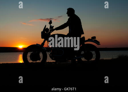 Motorbiker motorcyclist  in silhouette at sunset man on motorbike one hand on handlebar Stock Photo