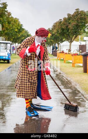 Red-nosed Sunny the Clown, (Sonny and Rainbow) wearing swimming flippers & sweeping puddles away with a brush. Heavy rains couldn't dampen the spirits of the visitors to this year's Southport Flower Show.  Umbrellas and wellies were the top fashion tip of the day. The largest independent flower show in England, is expecting thousands of visitors over the four day event.  Show Chairman and Trustee David Rose has employed road sweepers & clowns to keep the paths clear of any puddles so the elegant ladies can keep their feet dry. Stock Photo