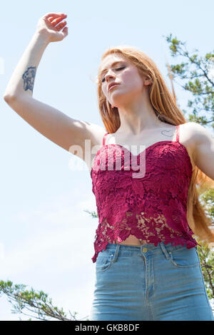 Beautiful red head in red top and skinny jeans, standing half length on mountain top with hand raised, looking down. Stock Photo