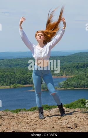 Beautiful red head in skinny jeans, black boots, and white top, on mountain top with wind in hair and a lake below. Stock Photo