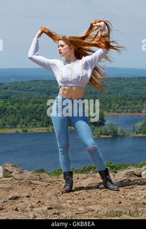 Beautiful red head in skinny jeans, black boots, and white top, on mountain top with wind in hair and a lake below. Stock Photo